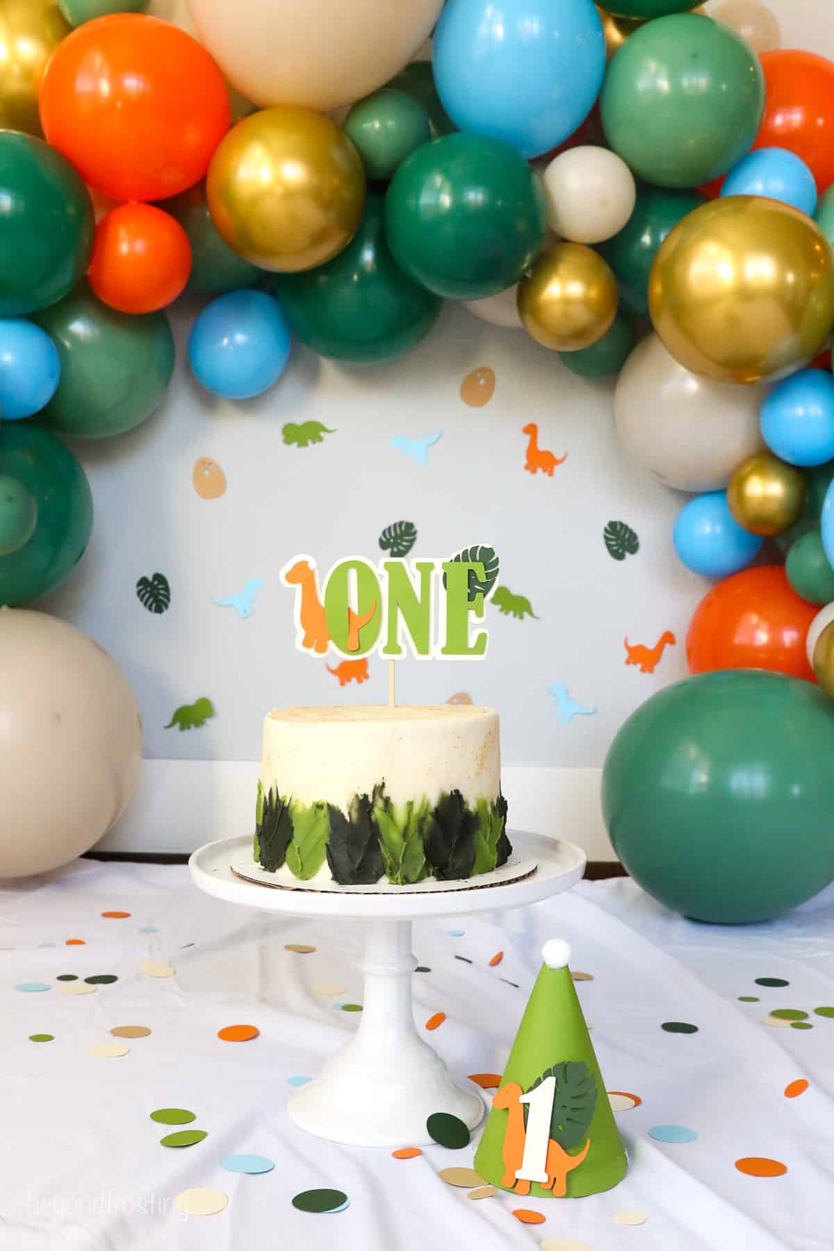 Full shot of a decorated dinosaur cake on a cake stand surrounded by confetti and a birthday hat, framed by a balloon arch in the background.