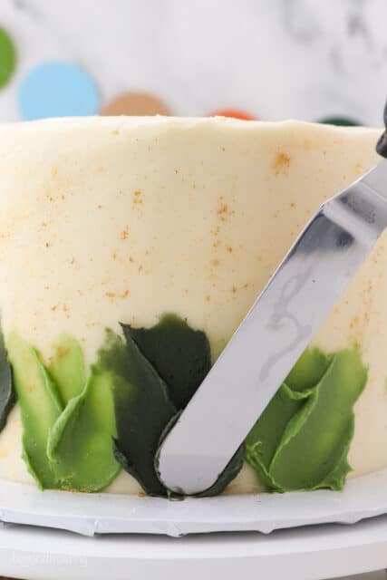 An offset spatula spreads shades of green frosting over the side of a gold-splattered cake to create a skirt.