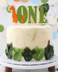 Dinosaur cake decorated with green frosting, topped with a dinosaur-themed "One" cake topper on a cake stand.