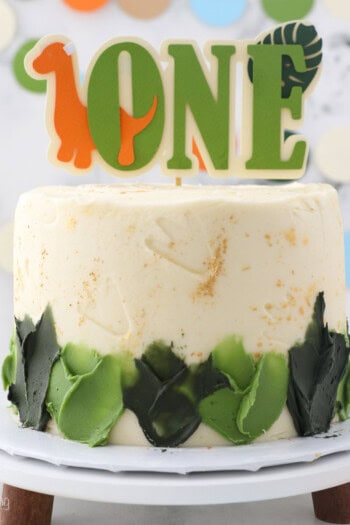 Dinosaur cake decorated with green frosting, topped with a dinosaur-themed "One" cake topper on a cake stand.