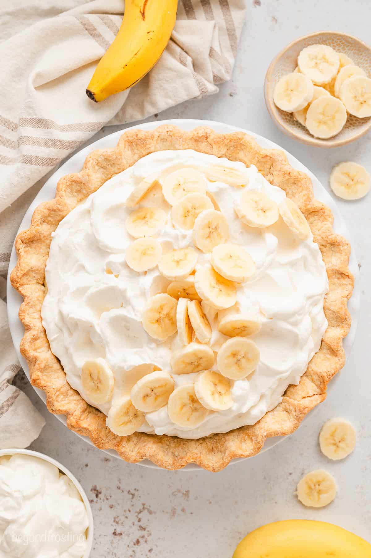 Overhead view of a whole banana cream pie garnished with banana slices, surrounded by pie ingredients.