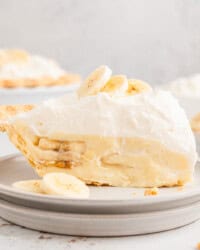 A slice of banana cream pie topped with banana slices on a white plate, with the rest of the pie in the background.