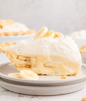 A slice of banana cream pie topped with banana slices on a white plate, with the rest of the pie in the background.