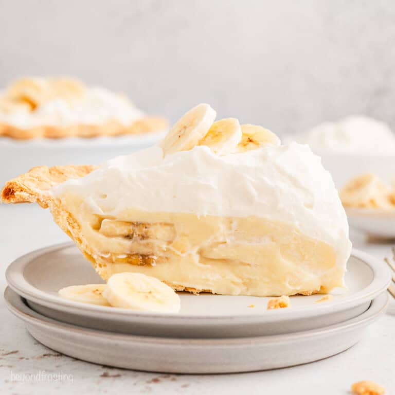 A slice of banana cream pie topped with banana slices on a white plate, with the rest of the pie in the background.