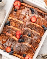 Overhead view of a baked brioche French toast casserole topped with powdered sugar and garnished with fresh strawberries and blueberries.