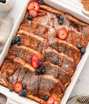 Overhead view of a baked brioche French toast casserole topped with powdered sugar and garnished with fresh strawberries and blueberries.