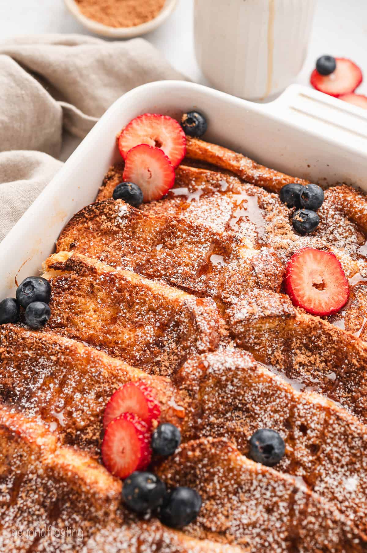 Close up of a baked brioche French toast casserole topped with powdered sugar and garnished with fresh strawberries and blueberries.
