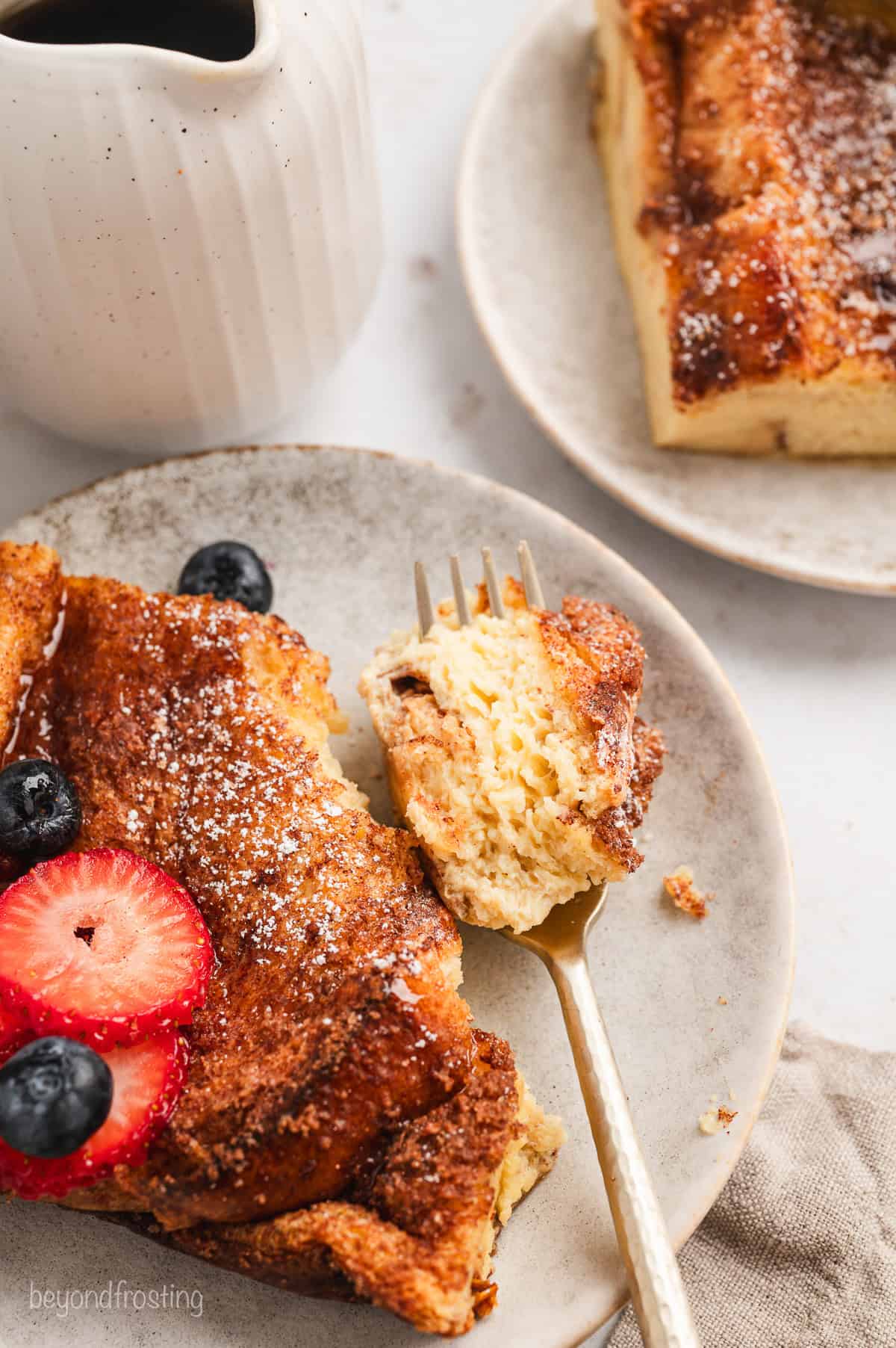 Overhead view of a forkful of French toast casserole next to a slice of French toast topped with berries on a plate, and a jug of maple syrup nearby.