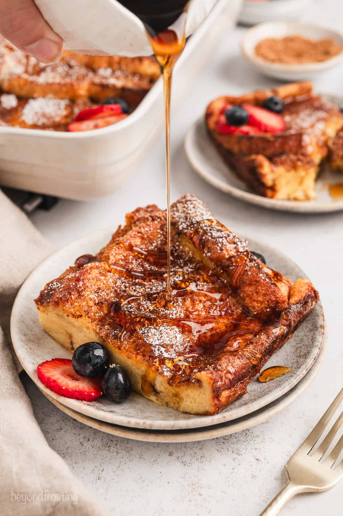 Maple syrup is drizzled from a jug over top of a slice of brioche French toast on a plate, with the casserole in the background.