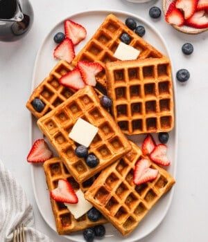 Overhead view of buttermilk waffles arranged on a platter, topped with butter and fresh strawberries and blueberries, next to bowls of toppings.