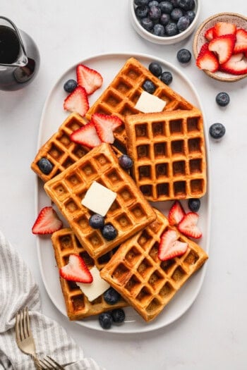 Overhead view of buttermilk waffles arranged on a platter, topped with butter and fresh strawberries and blueberries, next to bowls of toppings.