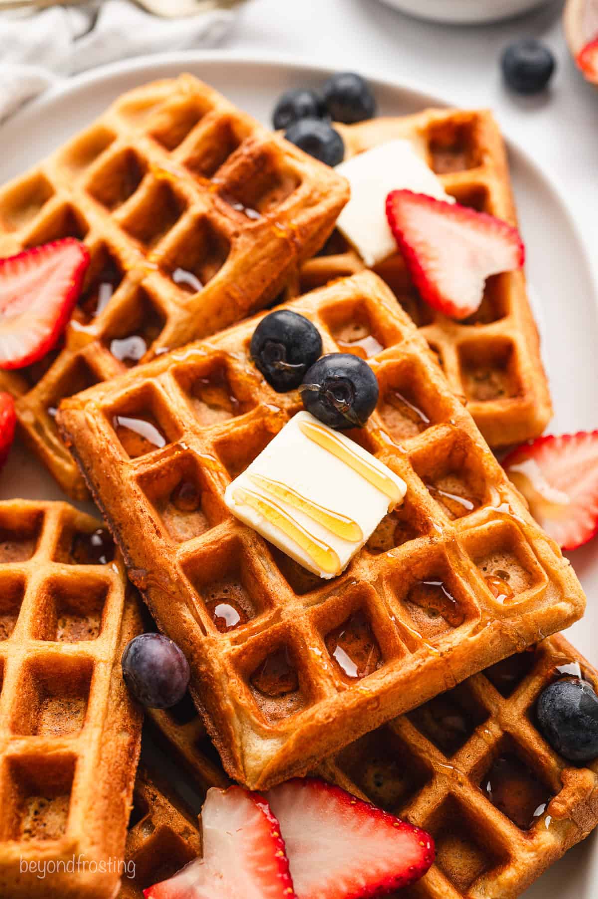 Close up of buttermilk waffles on a platter topped with maple syrup, butter, and fresh strawberries and blueberries.