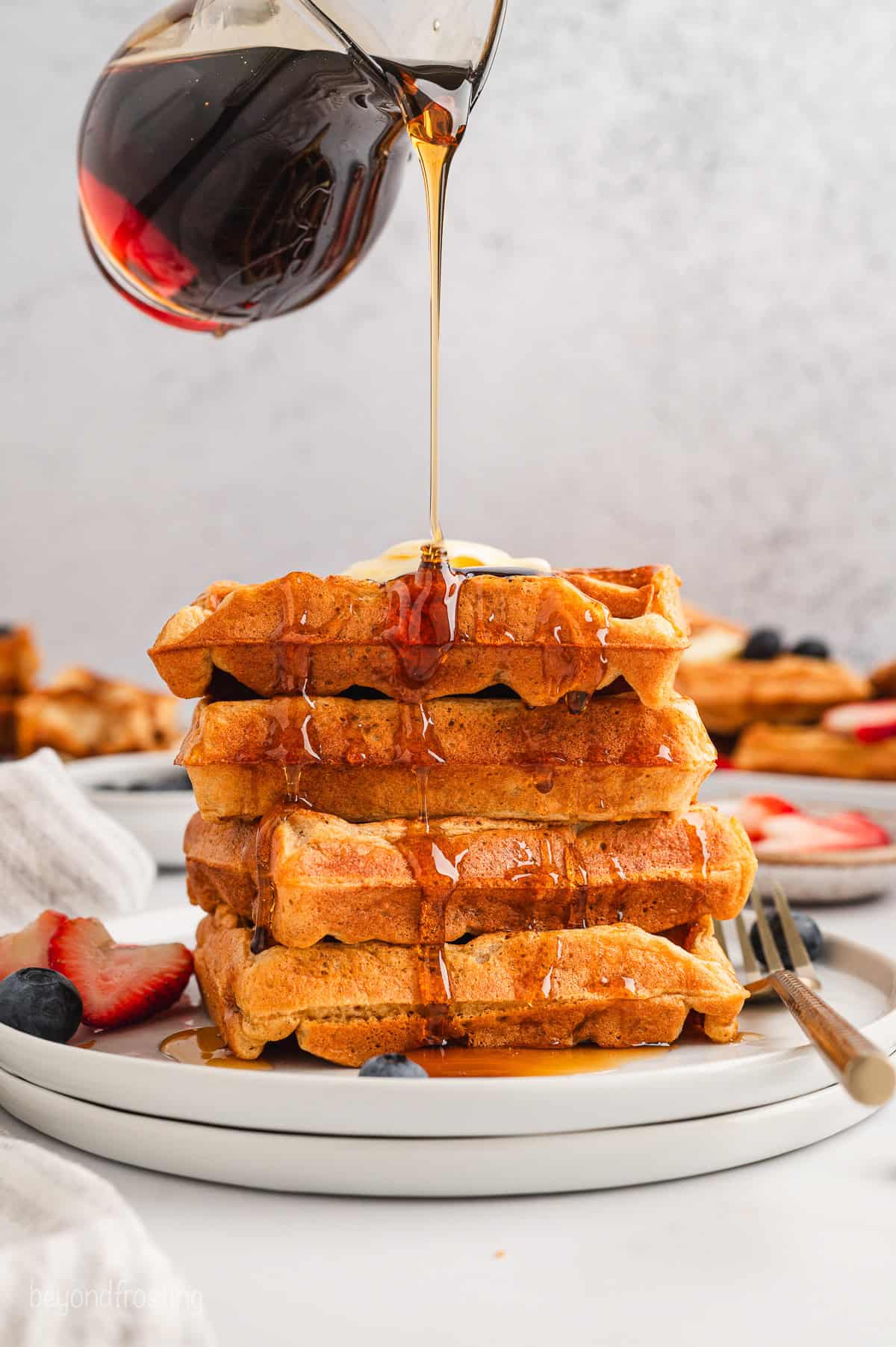 Maple syrup is drizzled from a jug over top of a stack of buttermilk waffles on a plate.
