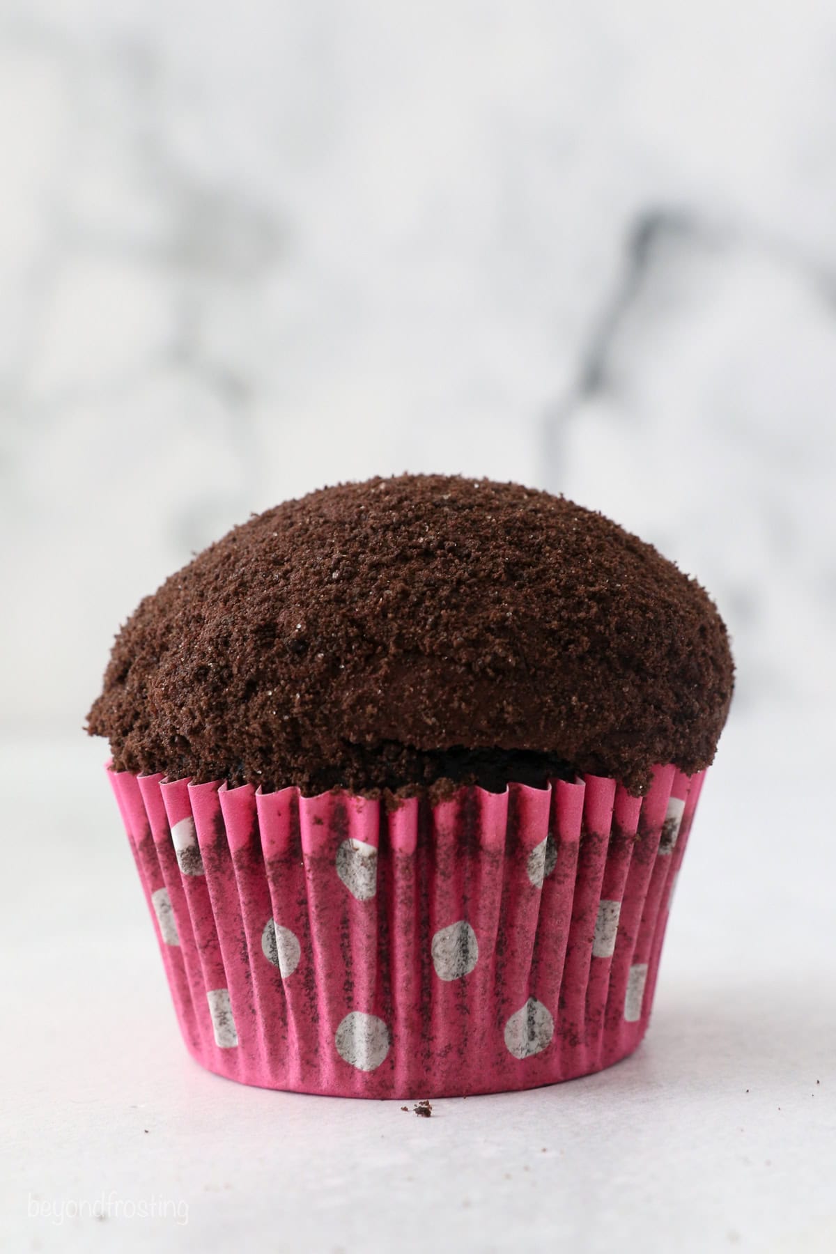 A frosted cupcake coated with Oreo crumbs inside a pink and white polka-dot cupcake liner.
