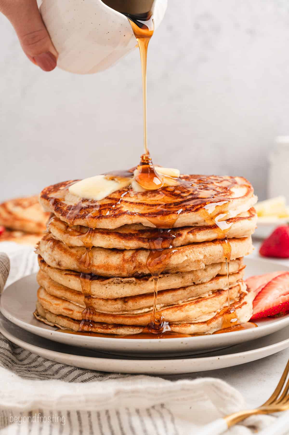 Maple syrup being poured from a small jug over a stack of buttermilk pancakes on a plate.