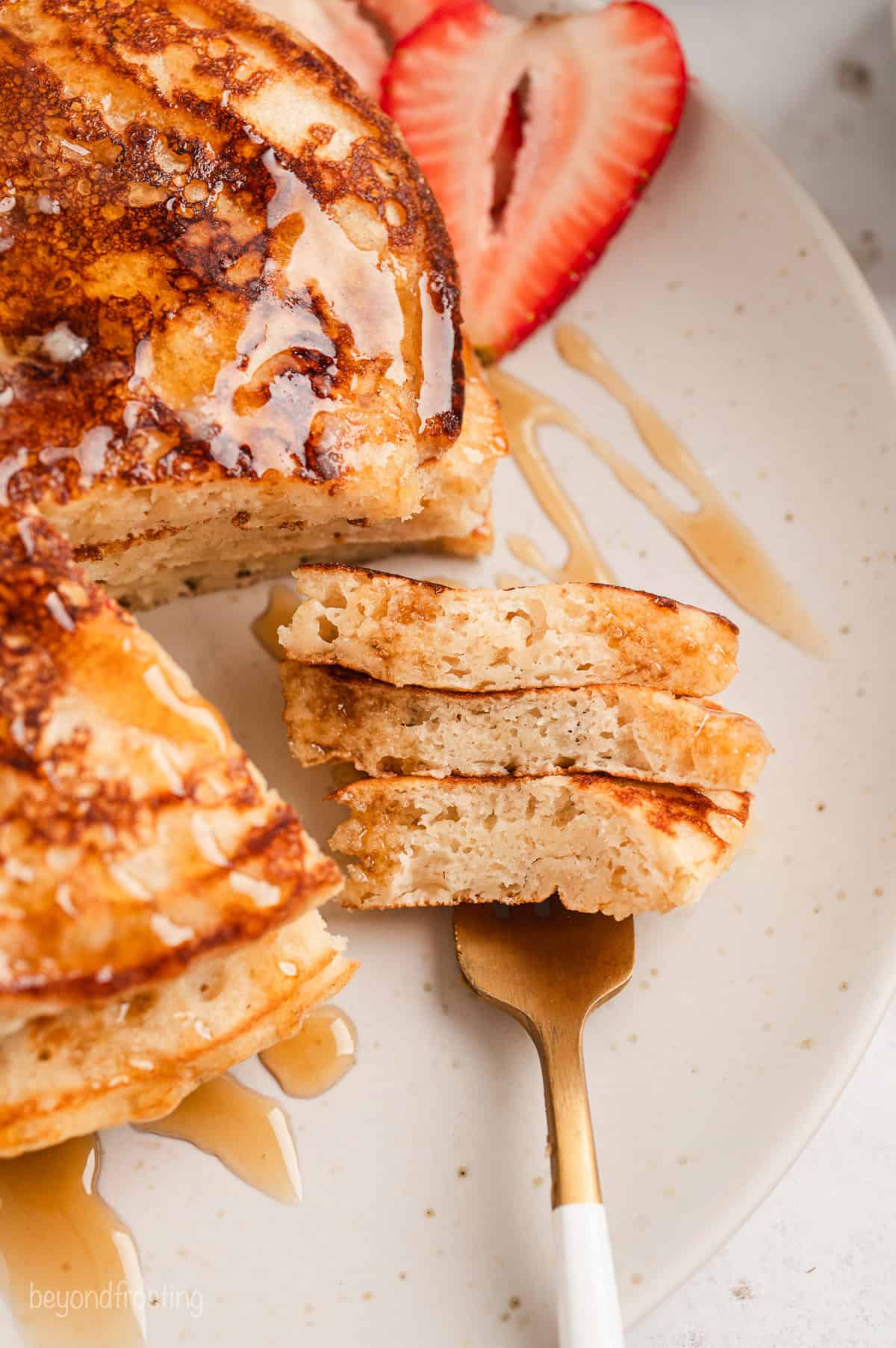 A forkful of buttermilk pancakes resting next to a stack of pancakes drizzled with maple syrup on a white plate.