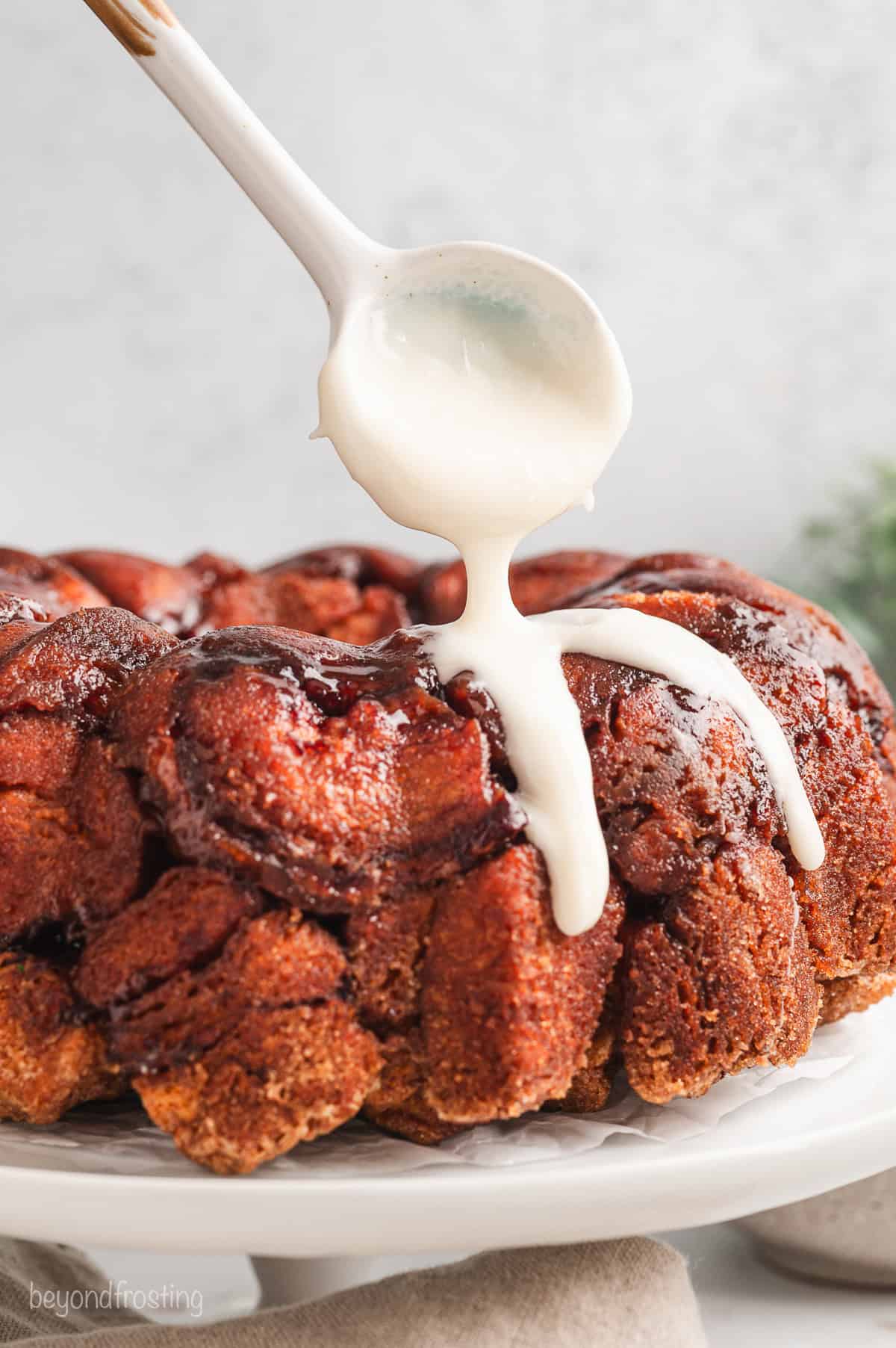 A spoon drizzling vanilla icing over cinnamon roll monkey bread on a cake stand.