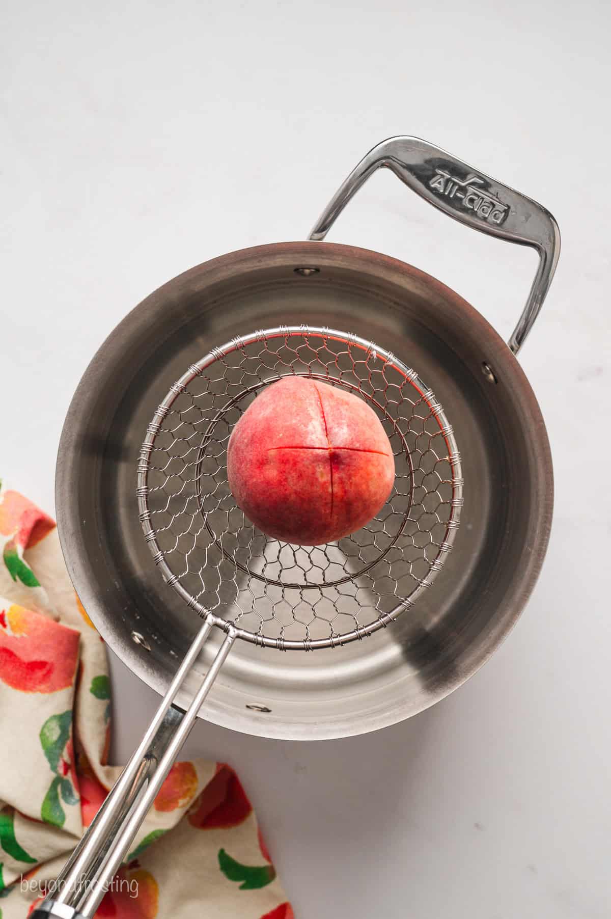 A blanched peach with an "X" scored in the top being lifted from a pot of boiling water with a slotted spoon.