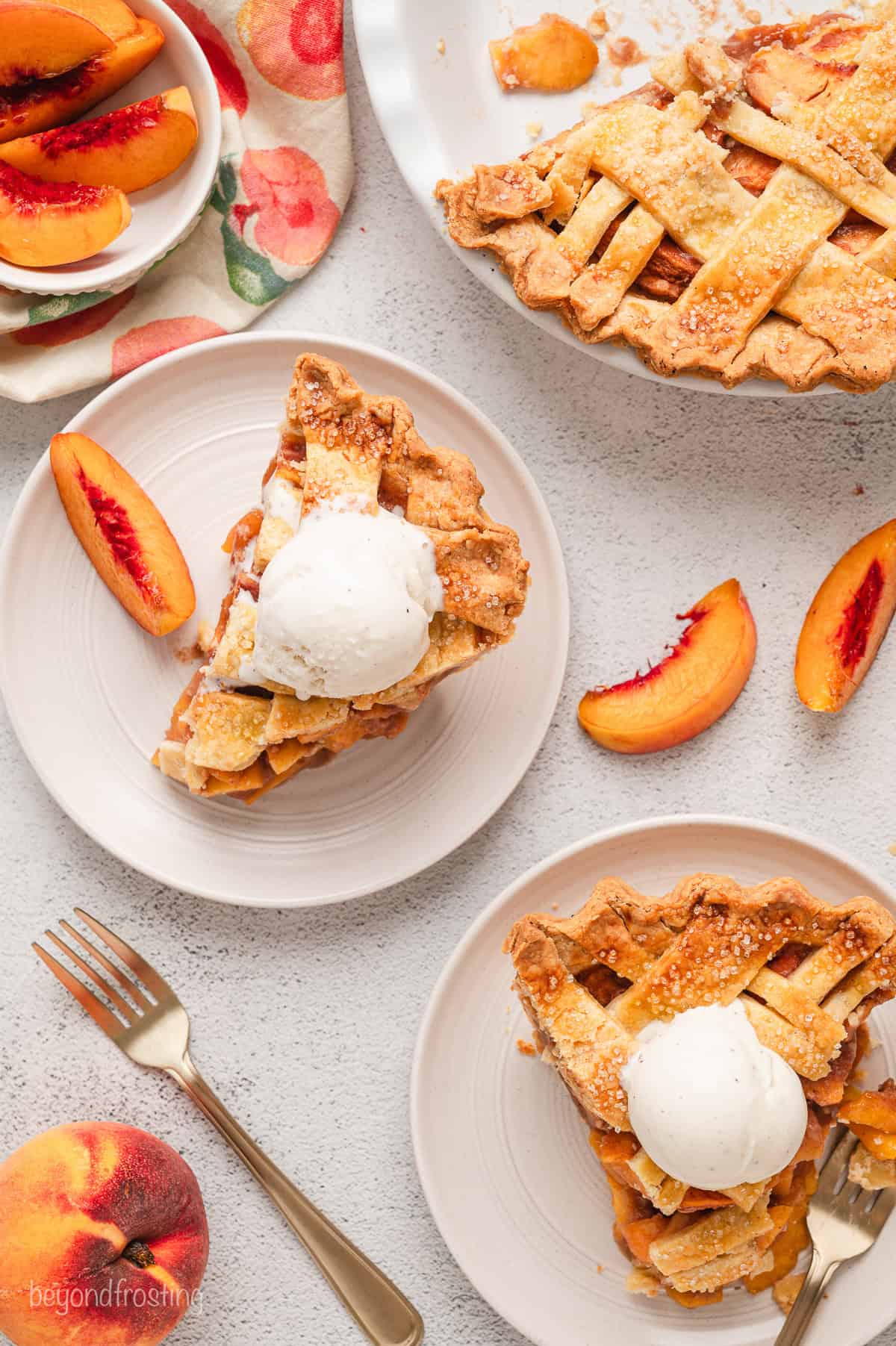 Overhead view of slices of peach pie topped with ice cream and served on plates, next to scattered peach slices and the rest of the pie.