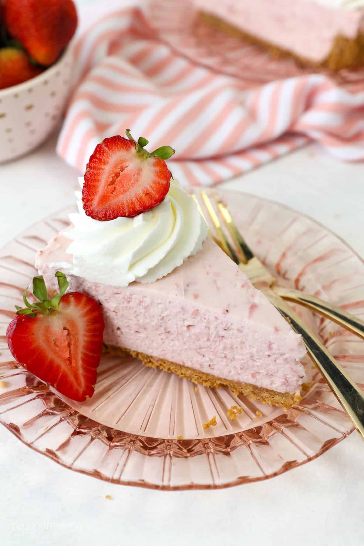 A slice of no-bake strawberry cheesecake on a pink glass plate, next to a fork and a fresh strawberry.