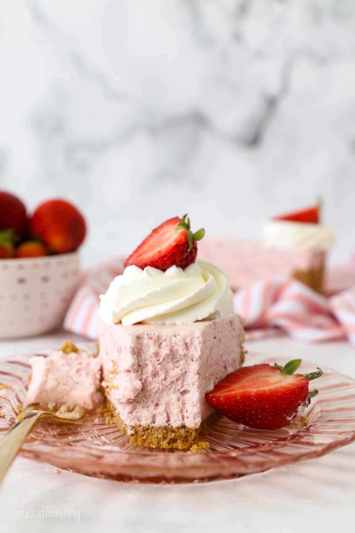 Head-on view of a slice of no-bake strawberry cheesecake on a pink glass plate, with a forkful missing from the end, next to a fork and a fresh strawberry.