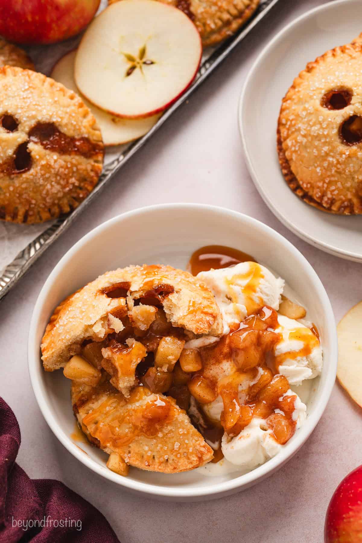 Overhead view of an apple hand pie served in a bowl with ice cream and caramel sauce, next to more hand pies on a plate and a baking sheet.