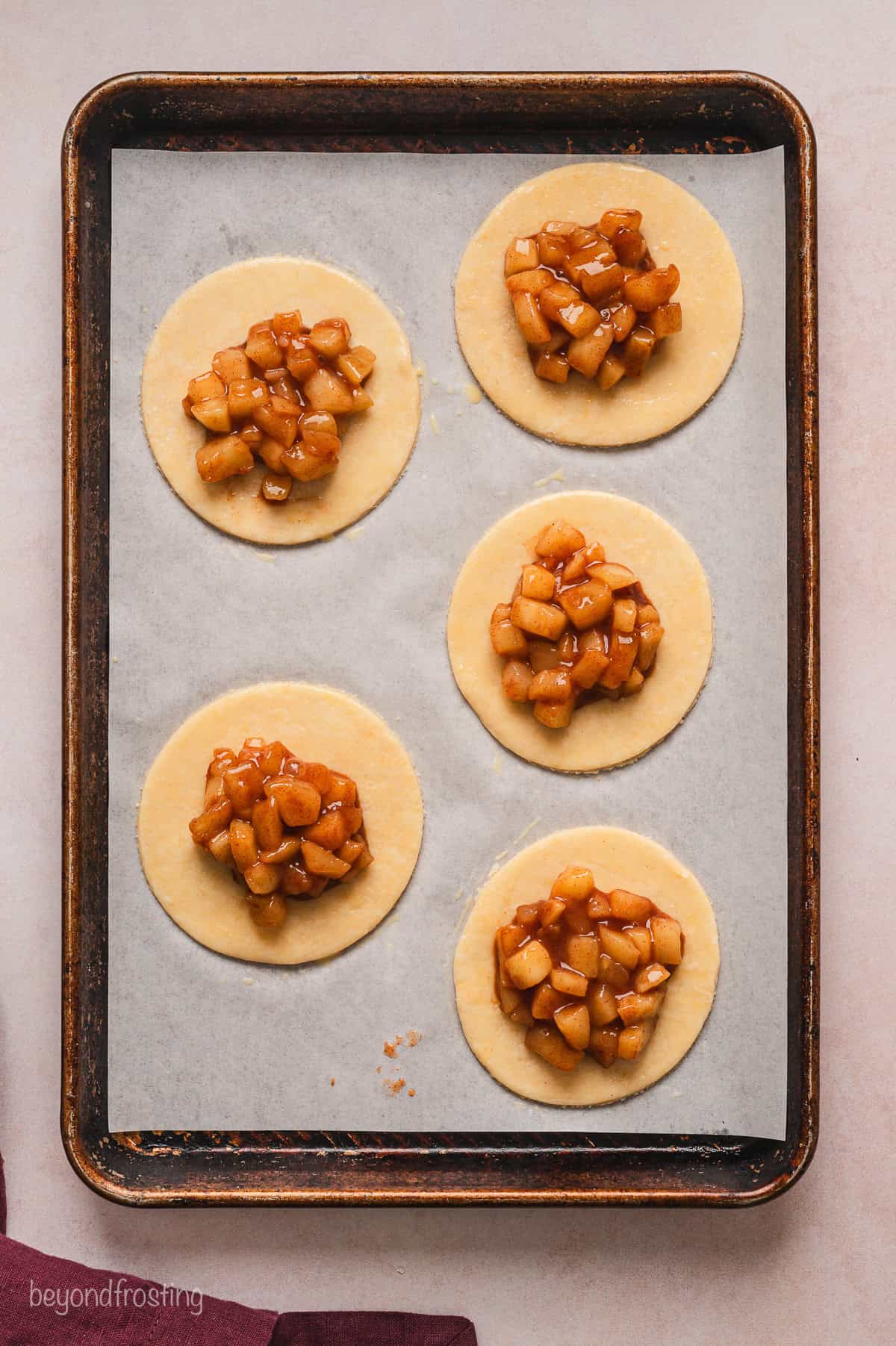 Five bottom pie crusts topped with apple filling on a parchment-lined baking sheet.