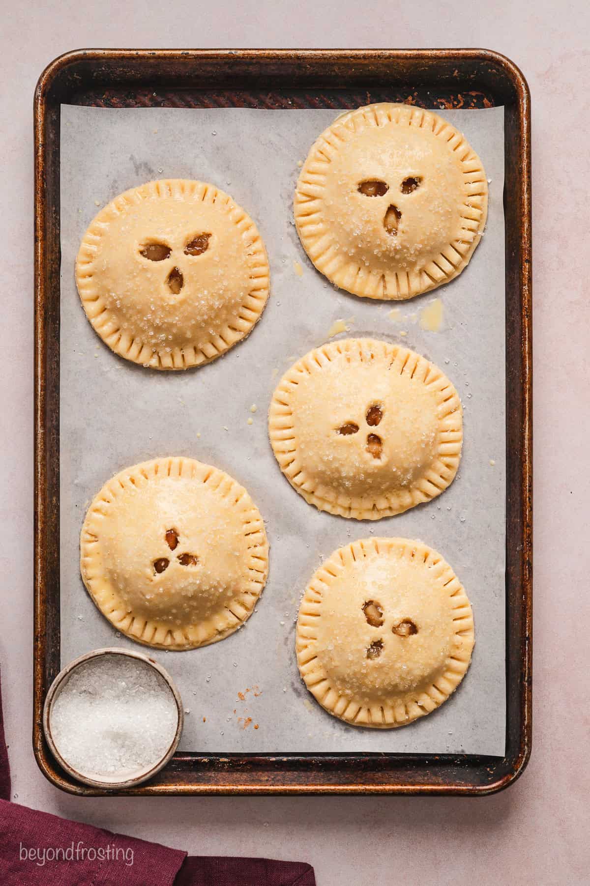 Five finished, unbaked hand pies on a parchment-lined baking sheet next to a small bowl of coarse sugar.