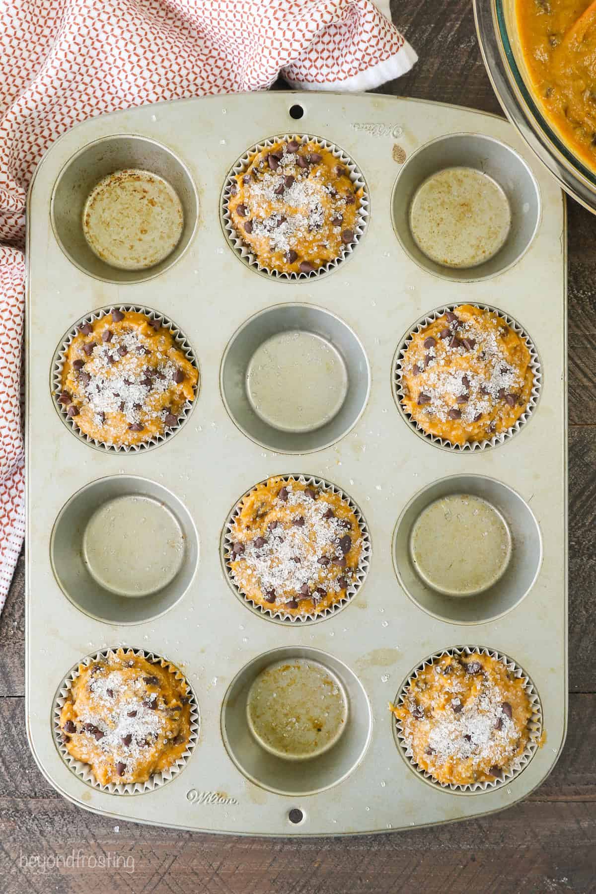 Overhead view of pumpkin chocolate chip muffin batter in alternating wells of a muffin tin, topped with sparkling sugar.