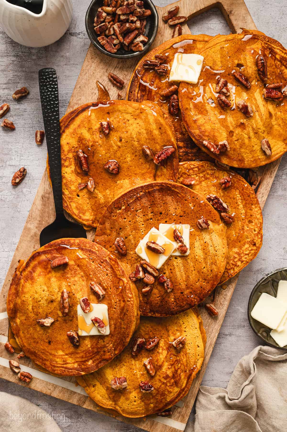 Overhead view of pumpkin pancakes on a wooden platter topped with pats of butter and scattered pecans.