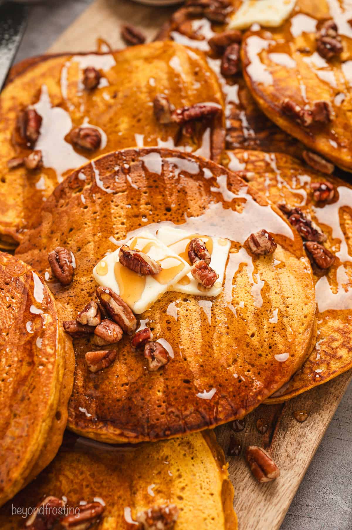 Close up view of pumpkin pancakes on a wooden platter topped with pats of butter and scattered pecans.