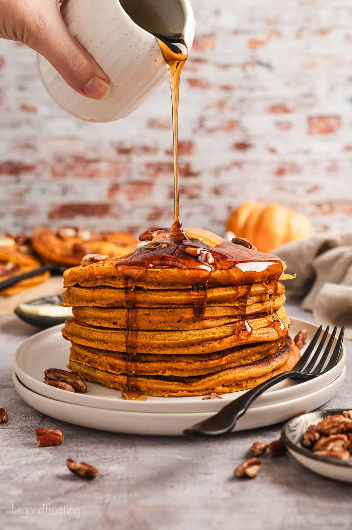 A hand drizzling maple syrup from a small jug over a stack of pumpkin pancakes garnished with pecans and butter.