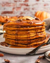 A tall stack of pumpkin pancakes on a plate, topped with pecans, butter, and maple syrup.