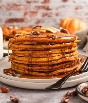 A tall stack of pumpkin pancakes on a plate, topped with pecans, butter, and maple syrup.