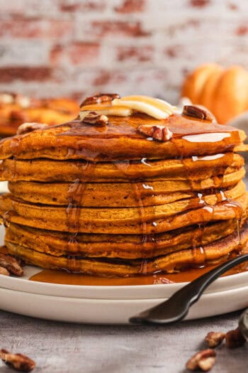 A tall stack of pumpkin pancakes on a plate, topped with pecans, butter, and maple syrup.