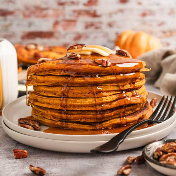 A tall stack of pumpkin pancakes on a plate, topped with pecans, butter, and maple syrup.