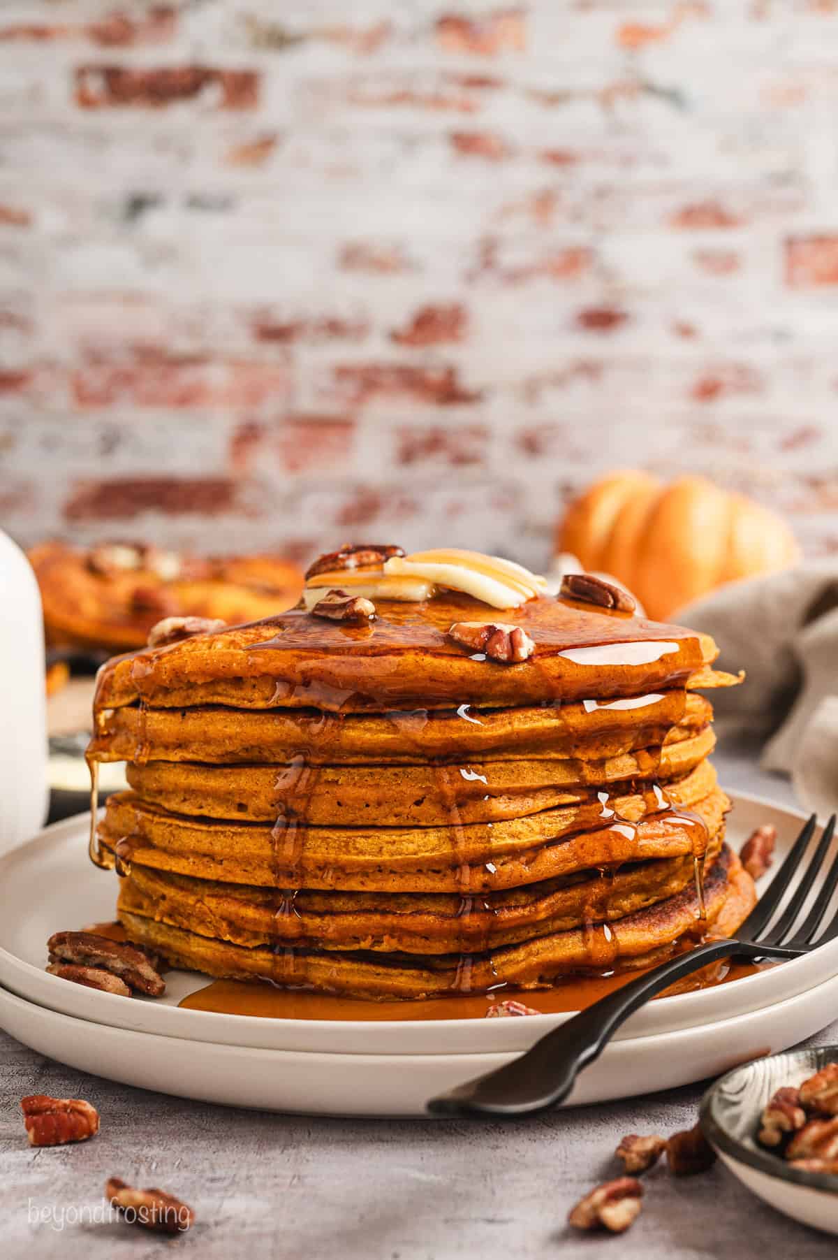 A tall stack of pumpkin pancakes on a plate, topped with pecans, butter, and maple syrup.