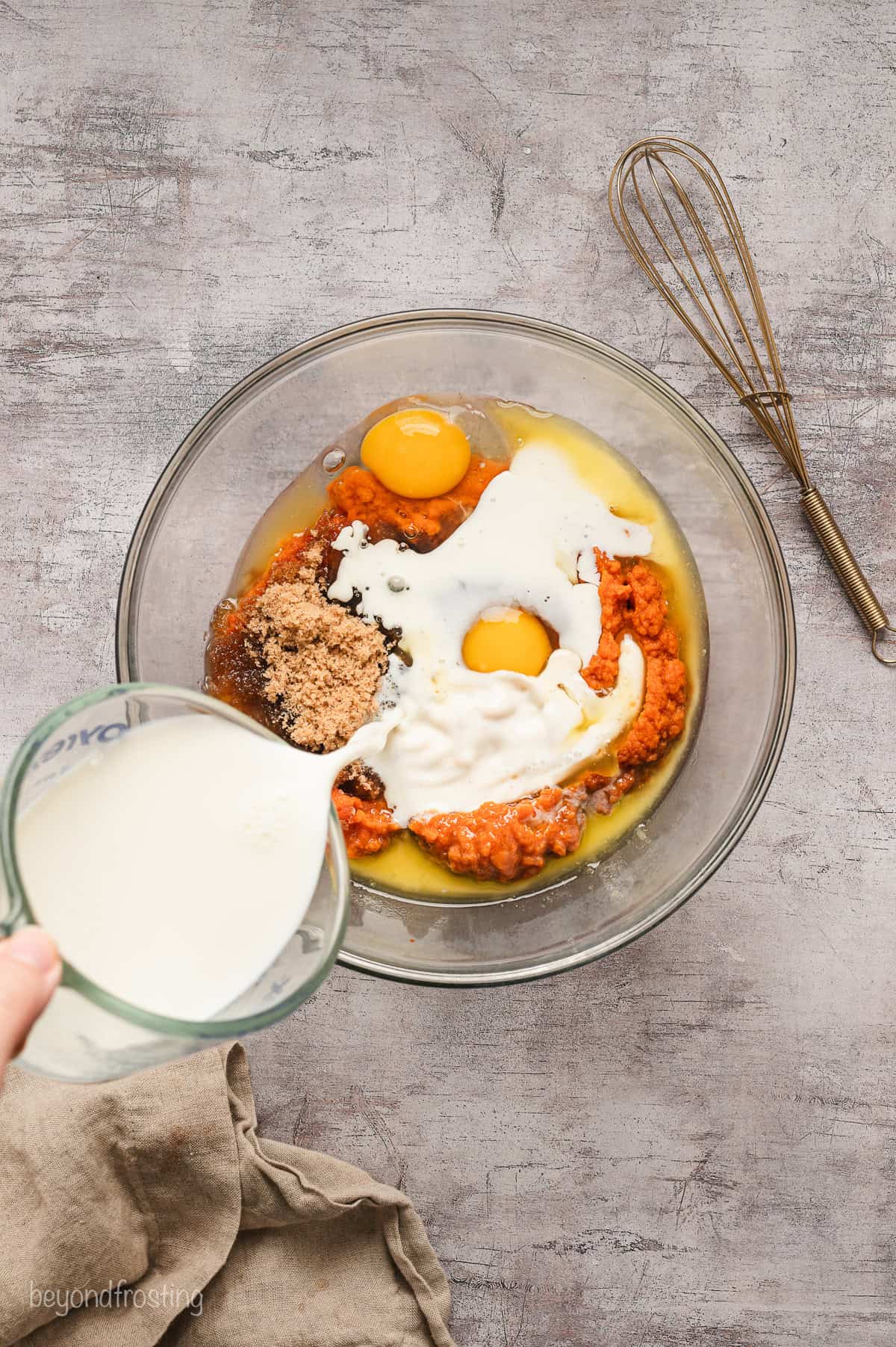 A hand pouring milk into a glass bowl with wet ingredients.