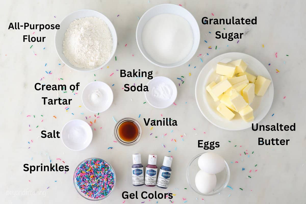 Ingredients for Unicorn cookies in small white bowls on a marble surface with sprinkles with labels over top