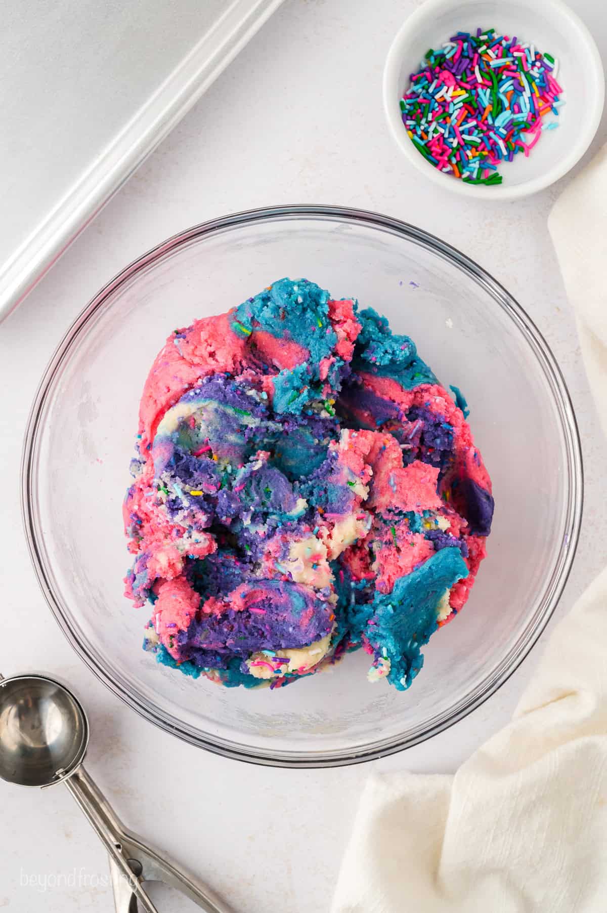 Overhead view of rainbow unicorn sugar cookie dough in a glass mixing bowl, next to a cookie scoop and a smaller bowl of sprinkles.