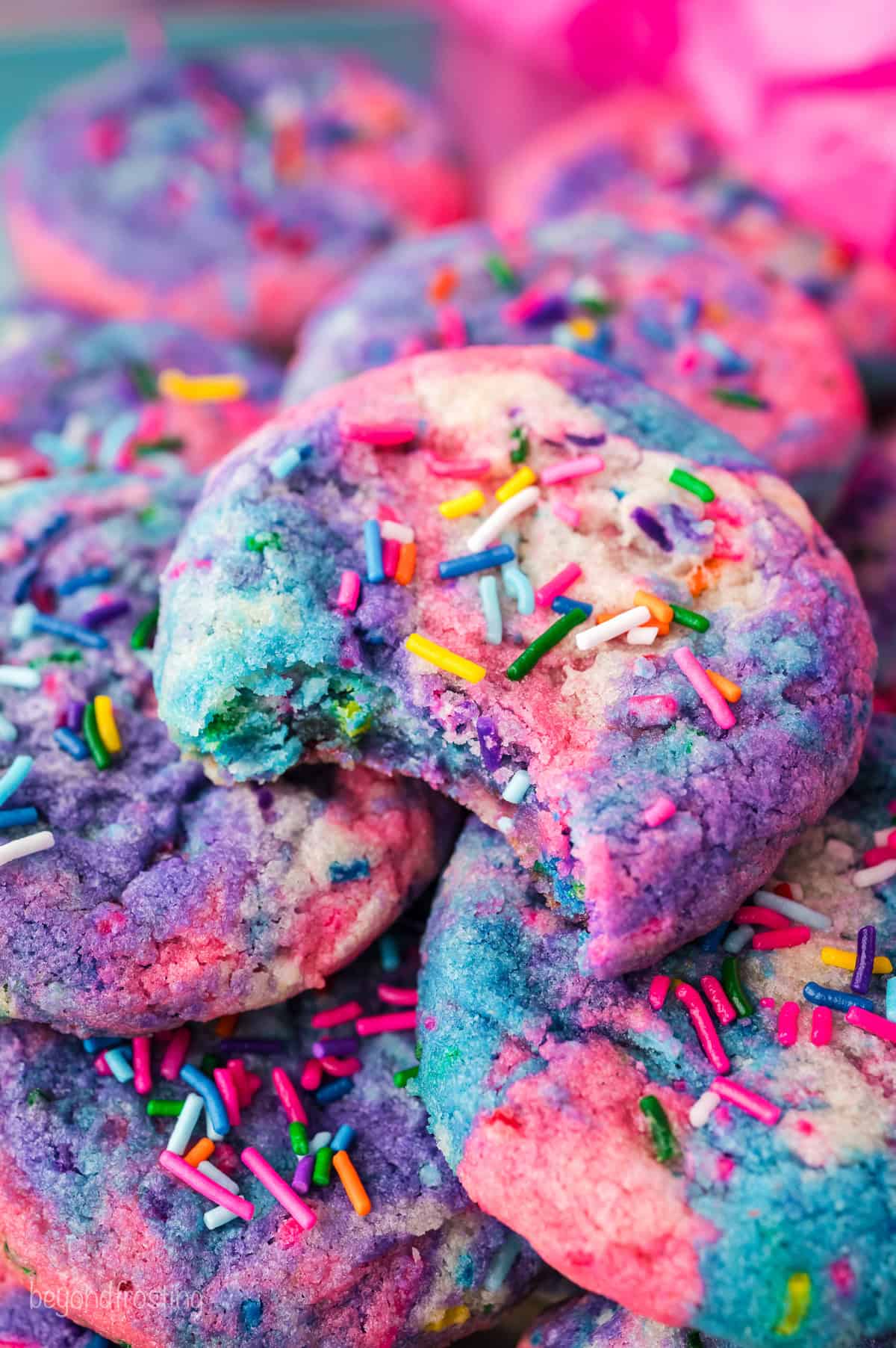 Close up of a unicorn cookie with a bite missing, resting on top of a pile of unicorn cookies decorated with rainbow sprinkles.