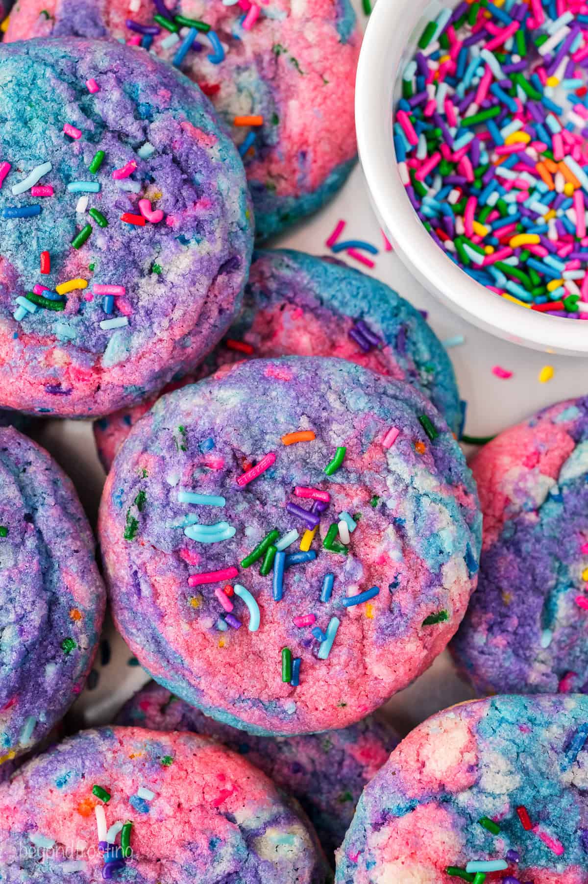 Overhead view of assorted unicorn cookies decorated with rainbow sprinkles, next to a bowl of sprinkles.