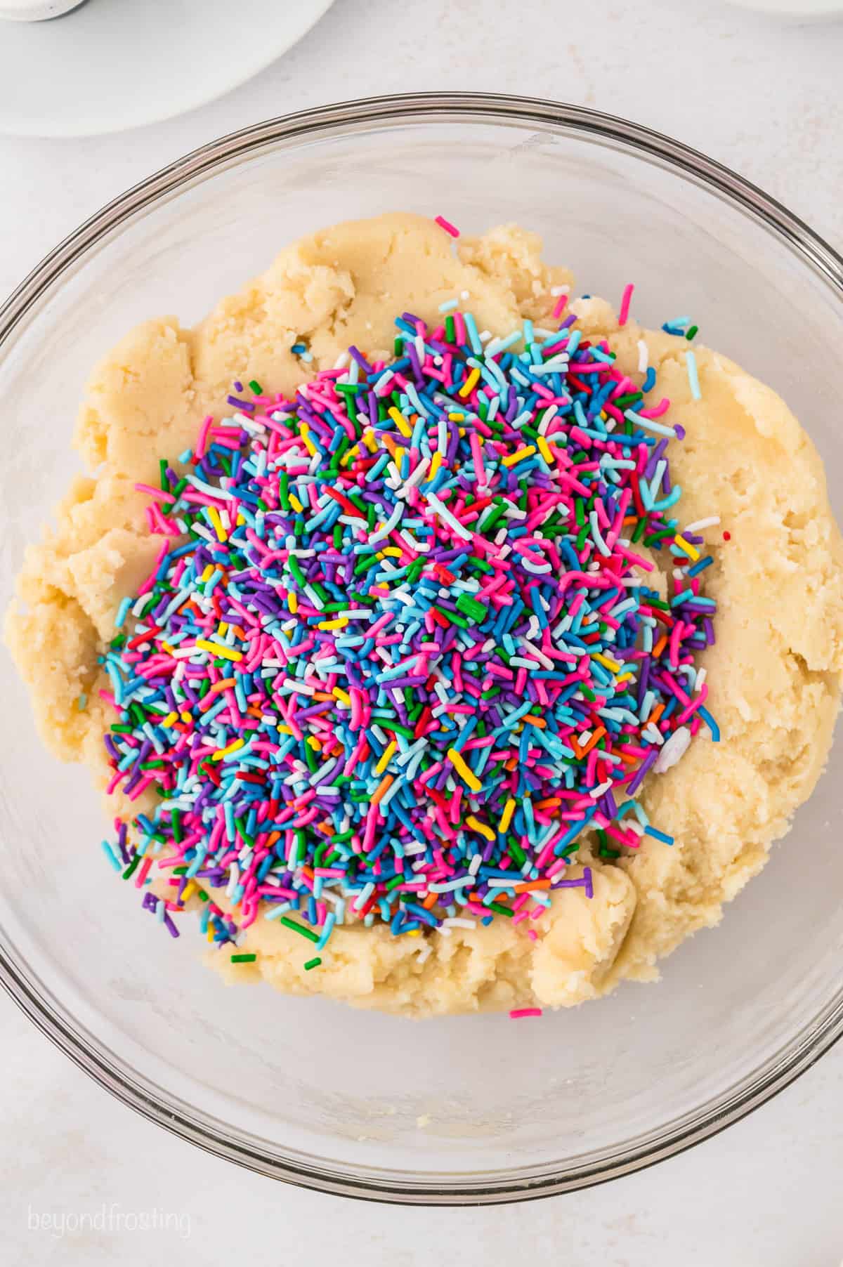 Rainbow sprinkles added to sugar cookie dough in a large glass mixing bowl.