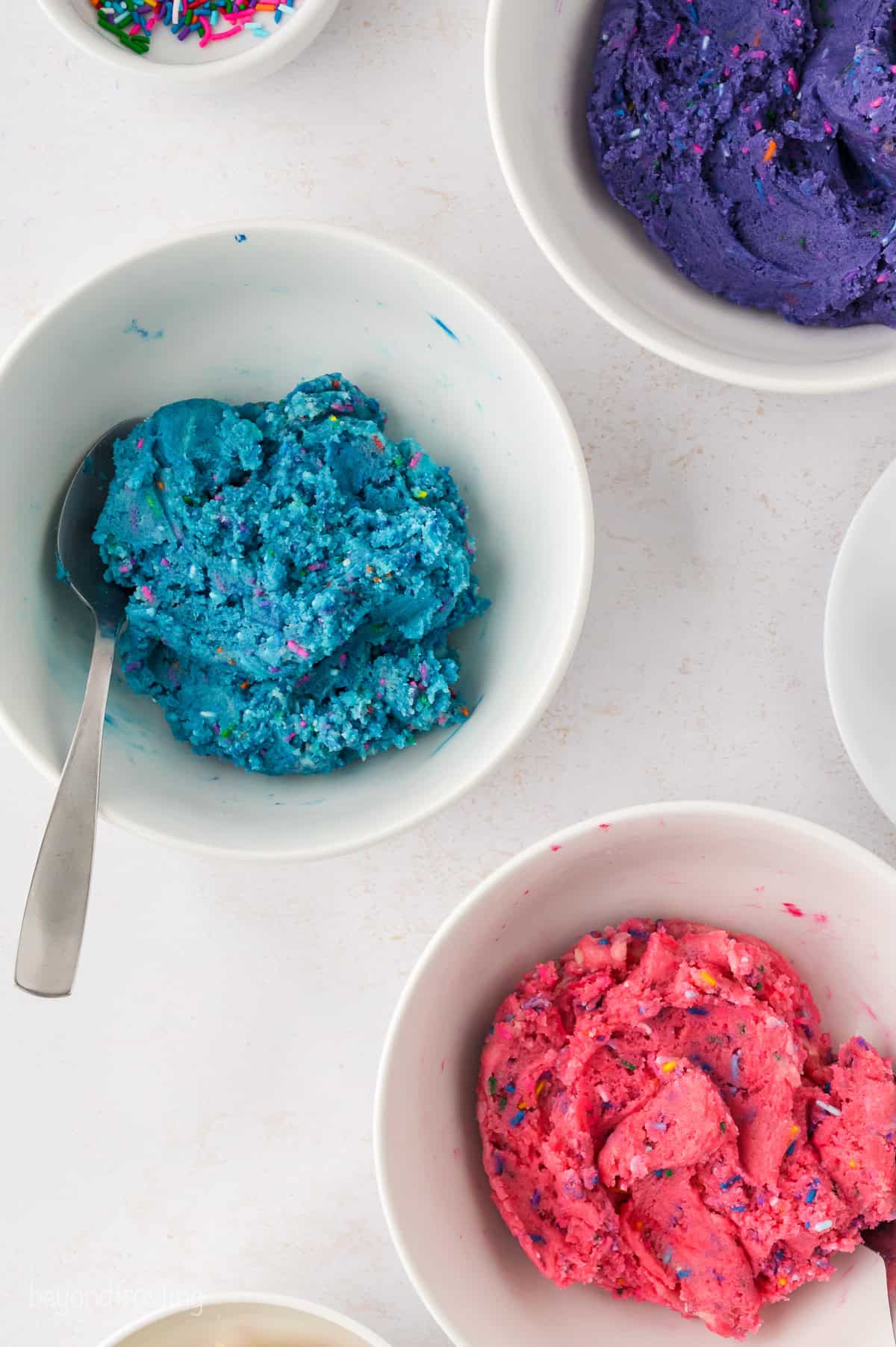 Overhead view of three white bowls filled with purple, blue, and pink colored unicorn cookie dough.