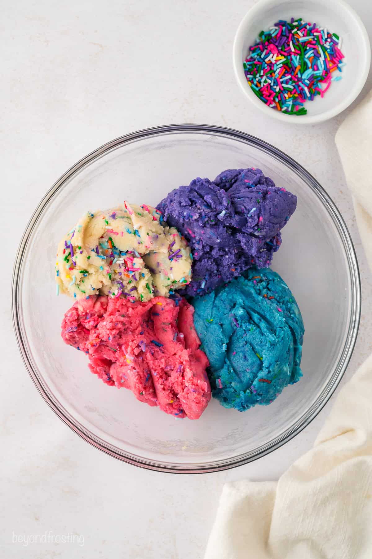 Portions of white, purple, pink, and blue sprinkle-filled sugar cookie dough added to a mixing bowl.