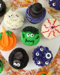 A cake stand with 7 different halloween cupcakes on top including a spider, monster themed, pumpkin, a mummy cupcake and a bleeding eye cupcake.