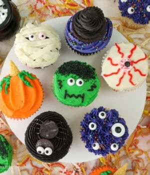 A cake stand with 7 different halloween cupcakes on top including a spider, monster themed, pumpkin, a mummy cupcake and a bleeding eye cupcake.