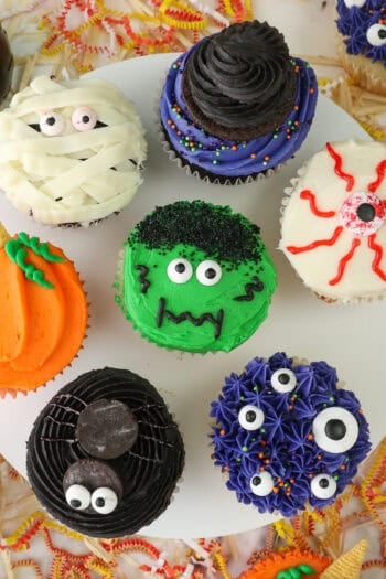 A cake stand with 7 different halloween cupcakes on top including a spider, monster themed, pumpkin, a mummy cupcake and a bleeding eye cupcake.