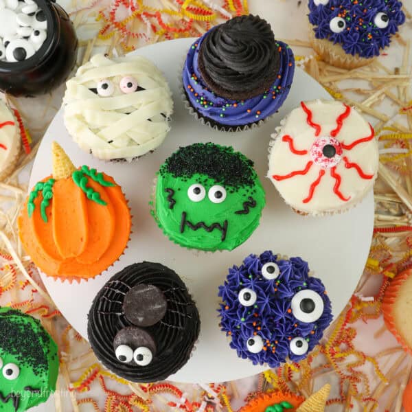 A cake stand with 7 different halloween cupcakes on top including a spider, monster themed, pumpkin, a mummy cupcake and a bleeding eye cupcake.