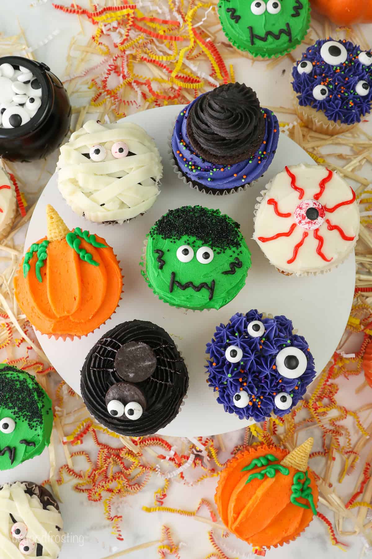 A cake stand with 7 different halloween cupcakes on top including a spider, monster themed, pumpkin, a mummy cupcake and a bleeding eye cupcake.